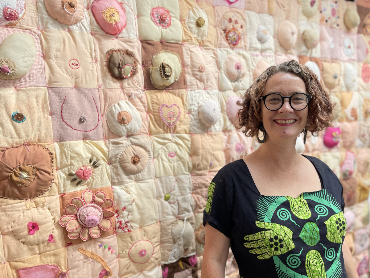 Artist Lois Blackburn, in front of her collaborative artwork Comfort. Art quilt made of 180 squares, each featuring an embroidered, knitted or crocheted breast. In many shades of pinks and browns, reflecting every size and shape of breast.
