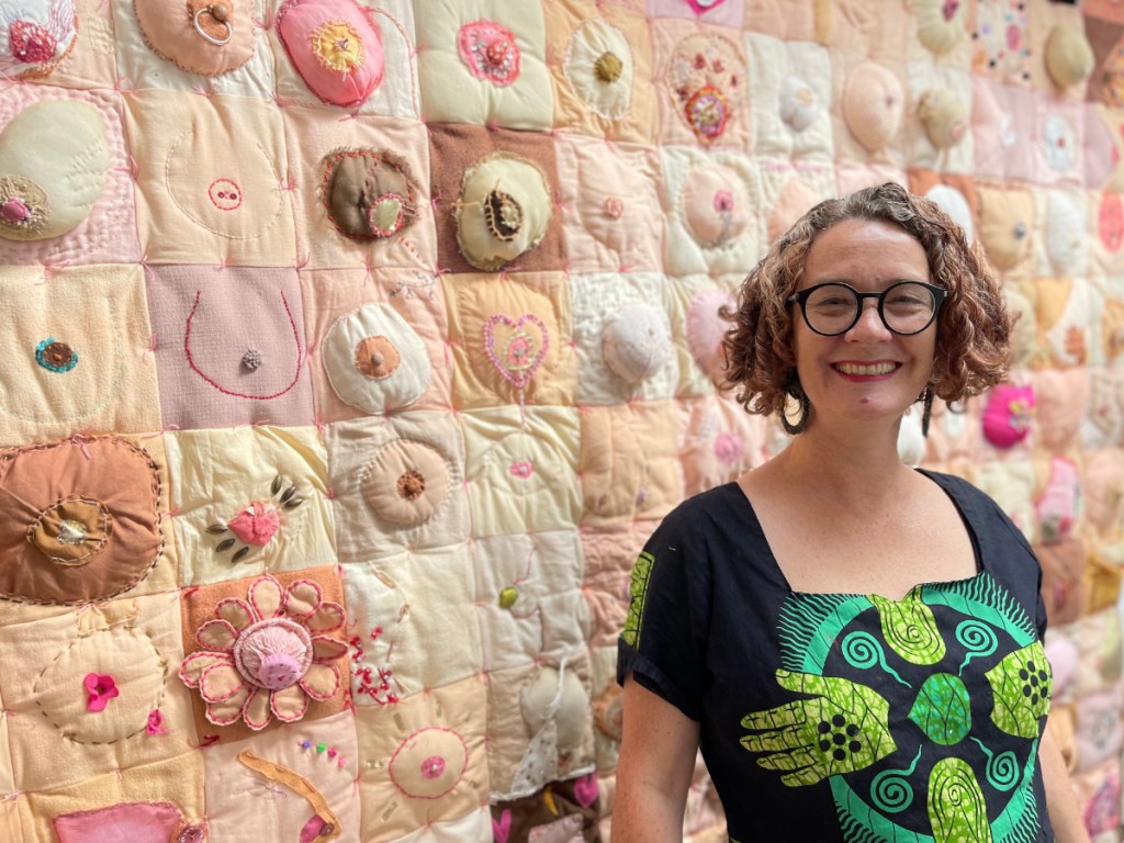 Artist Lois Blackburn, in front of her collaborative artwork Comfort. Art quilt made of 180 squares, each featuring an embroidered, knitted or crocheted breast. In many shades of pinks and browns, reflecting every size and shape of breast.
