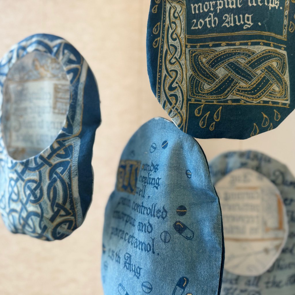 Close up of 4 stoma bag covers, hand made from reused cotton fabric. Each decorated with different designs, inspired by Celtic knots and illuminated manuscripts. In various shades of blue and cold.