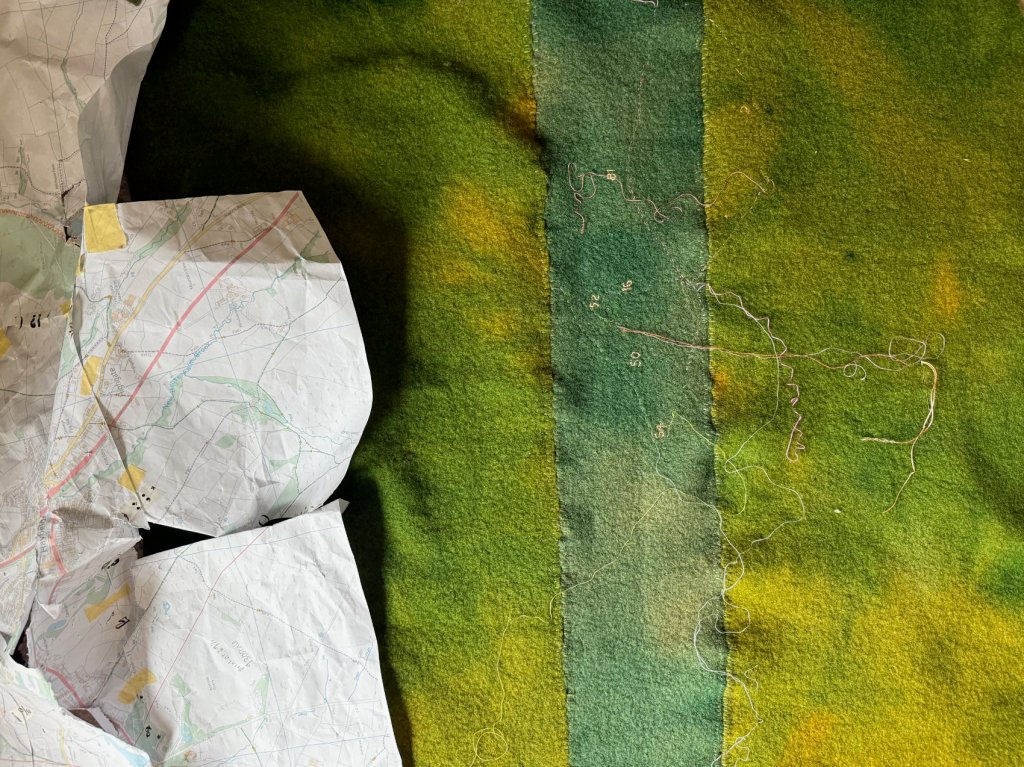 Printed maps showing footpaths, lying on top of stitched pieces of green dyed woollen blankets.